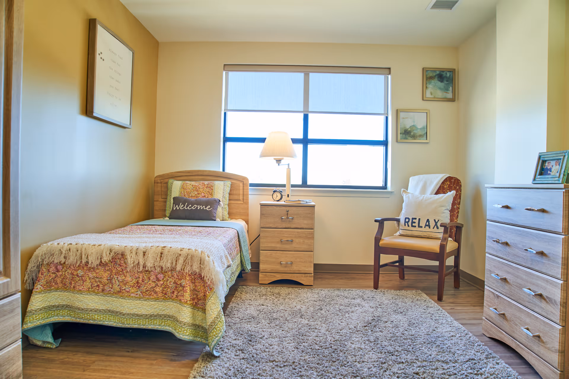Sunlit bedroom with bed, nightstand, chair, dresser, and window view.