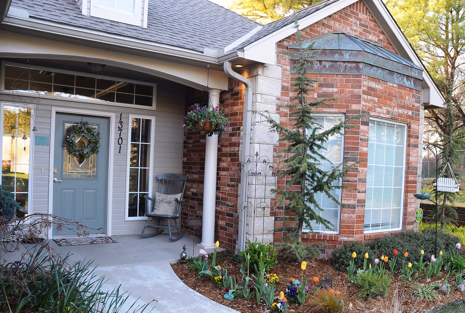 Front entrance of a senior living facility with a light blue door adorned with a wreath, a rocking chair with a cushion on the porch, and a garden with colorful flowers and small trees in front of a brick and stone exterior wall.
