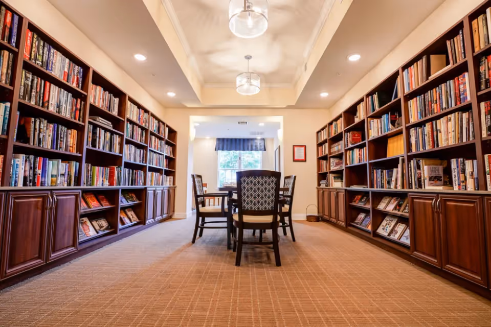 A cozy library/reading room with bookshelves lining both walls, a table and chairs in the center, and a window at the far end.