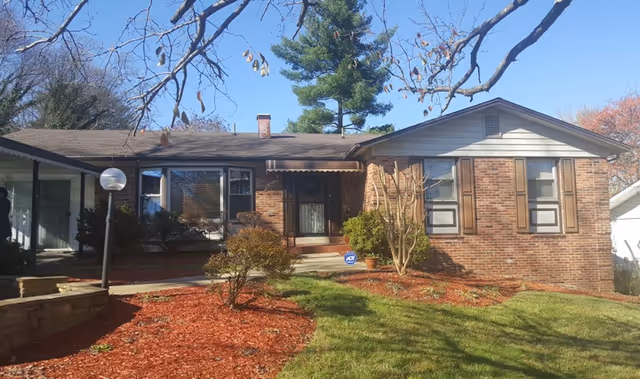 Single-story brick senior living facility front with lawn, shrubs, and leafless trees.