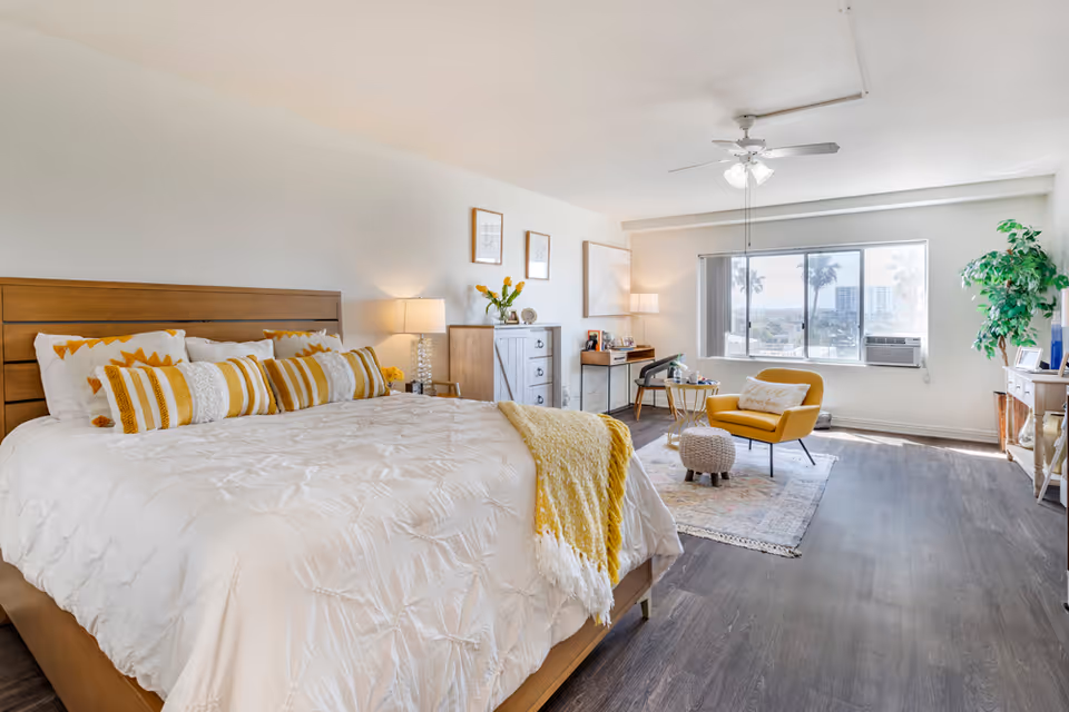 Bright, modern bedroom with a large bed, yellow accents, and seating area near windows.