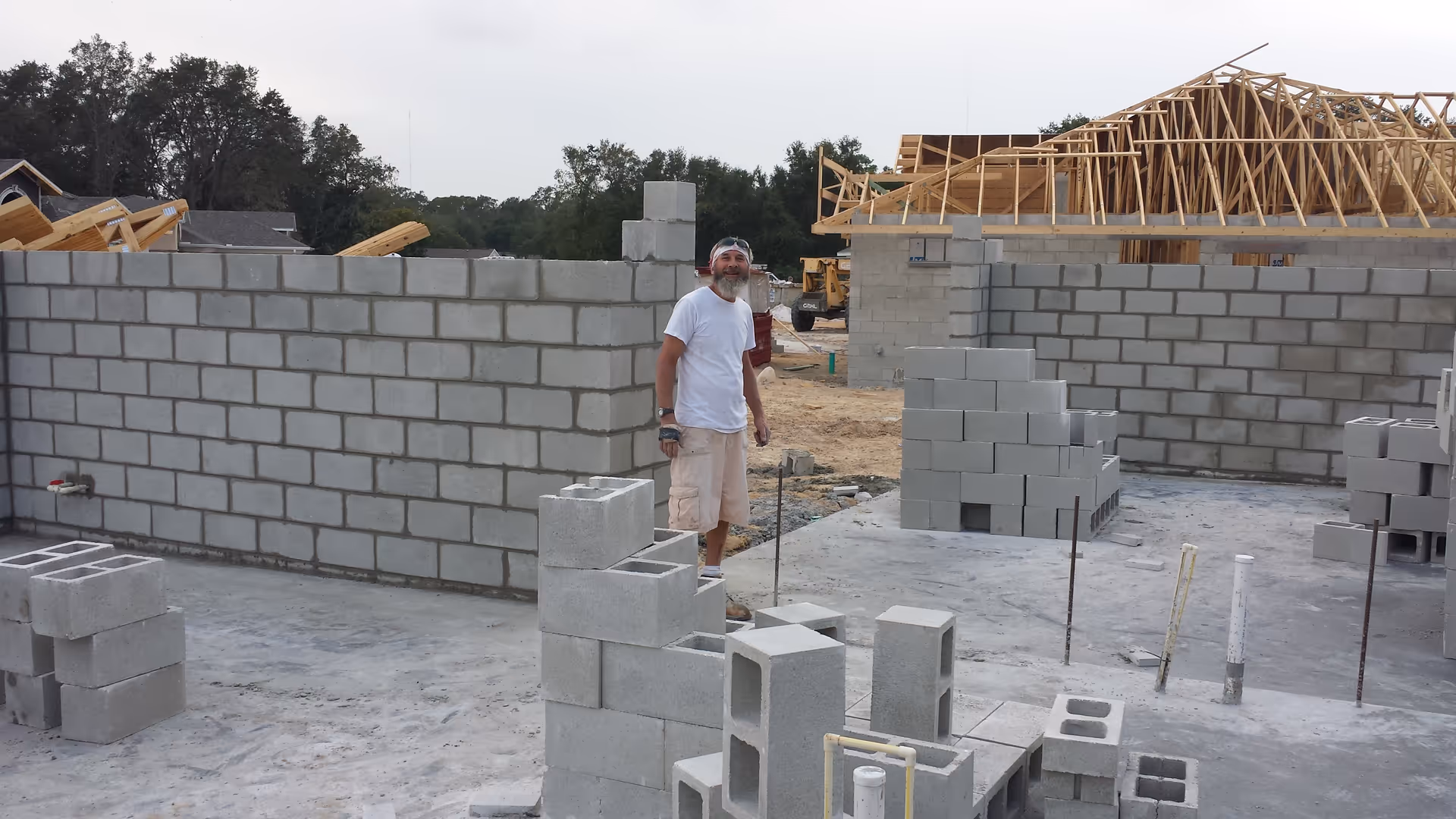 Construction site with a man standing near unfinished cinder-block walls and wooden roof framing.