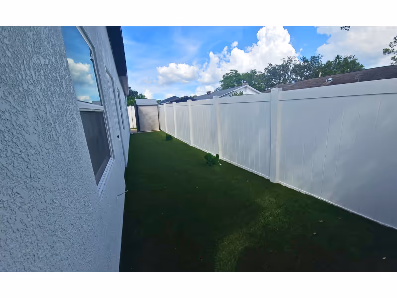 Narrow outdoor yard beside a stucco house, bordered by a tall white privacy fence.