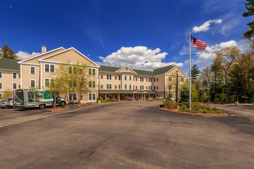 Exterior view of Bellamy Fields Assisted Living facility showing a large three-story beige building with multiple windows, a covered entrance, a parking area with a vehicle, an American flag on a flagpole, and trees surrounding the property under a partly cloudy blue sky.