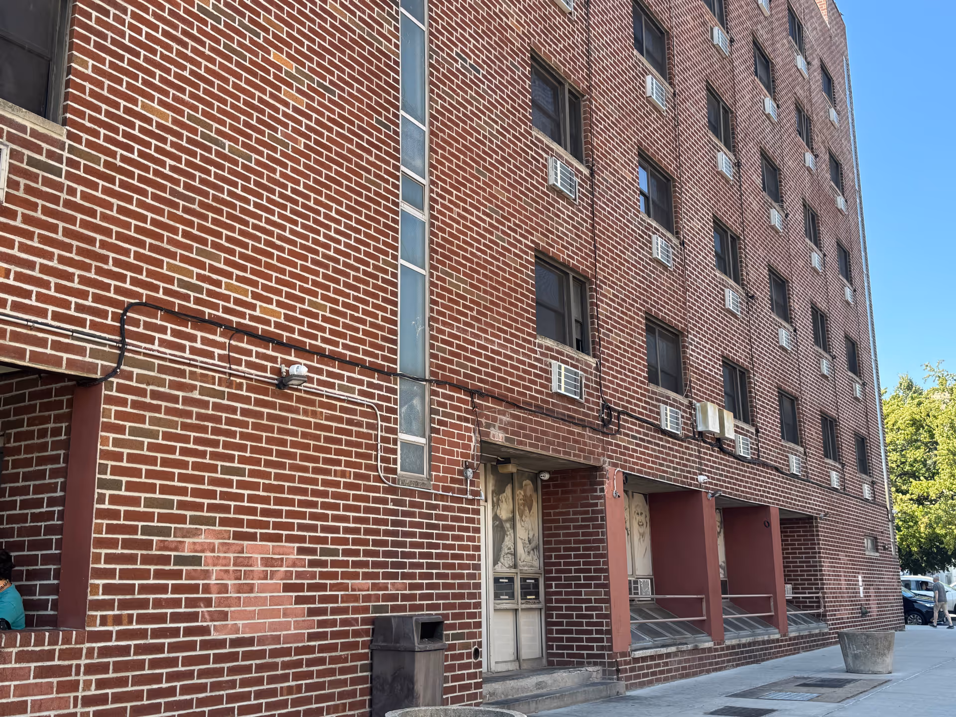 Red brick multi-story residential building with rows of windows, window air conditioners, and a street-level entrance.