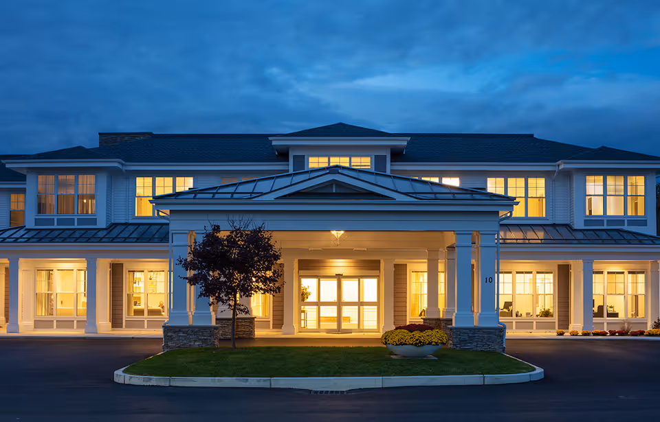 Front entrance of a two-story senior living residence at dusk with a lit covered portico and glowing windows.