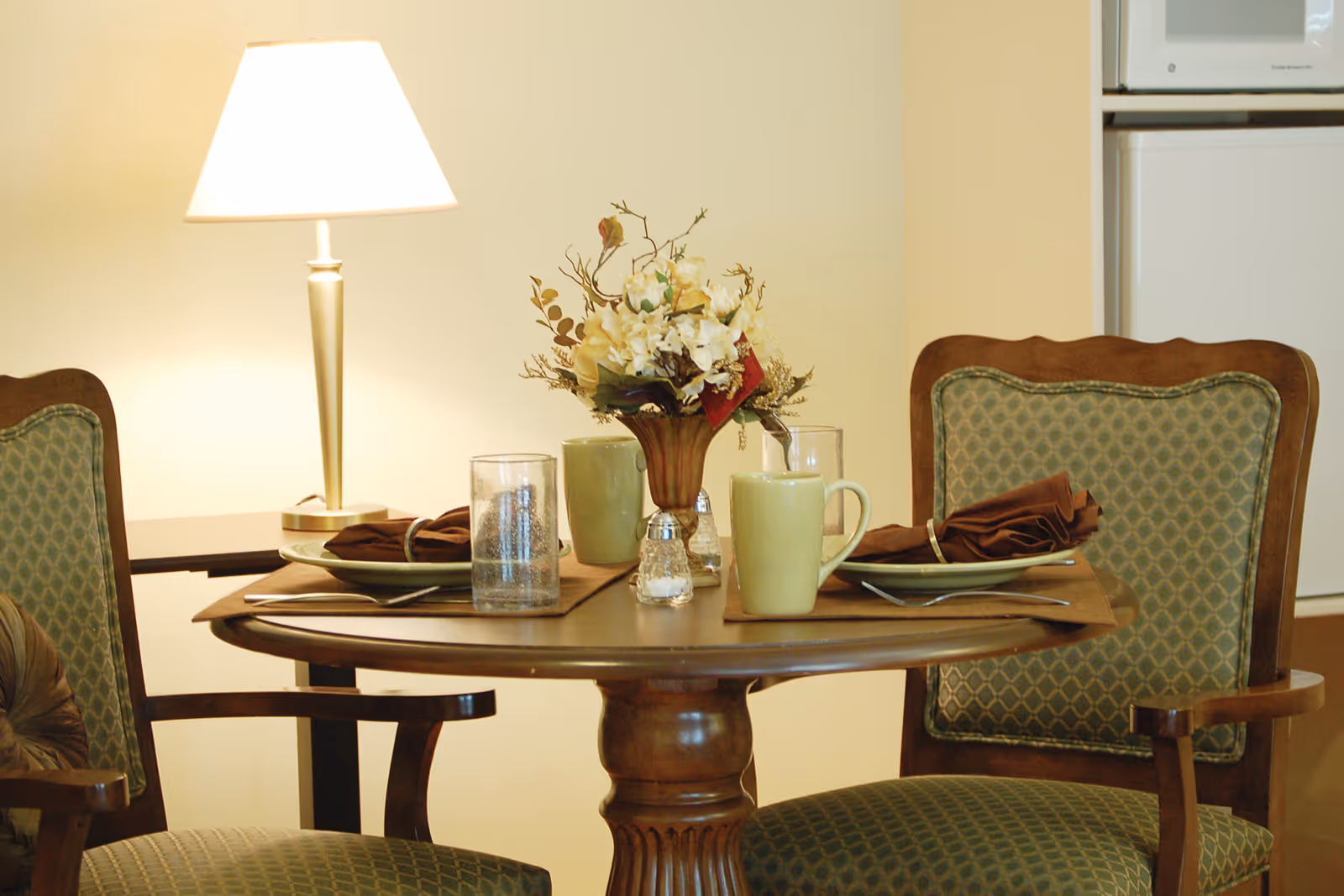 A dining table set with plates, cups, flowers, and a lamp in a cozy dining area.