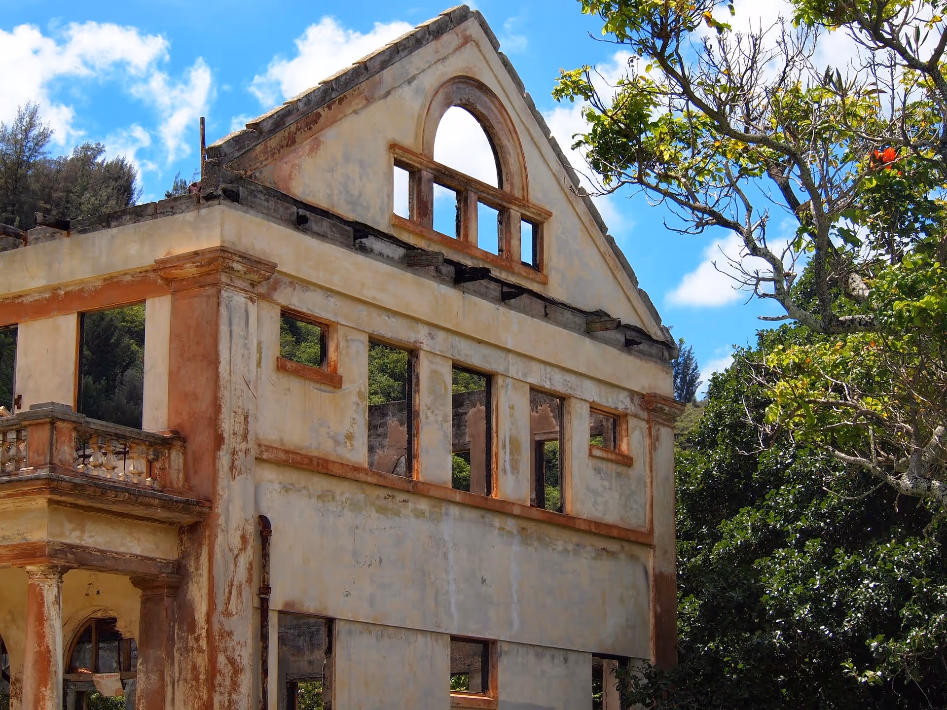 Ruined two-story building facade with broken windows and exposed framework beside leafy trees.