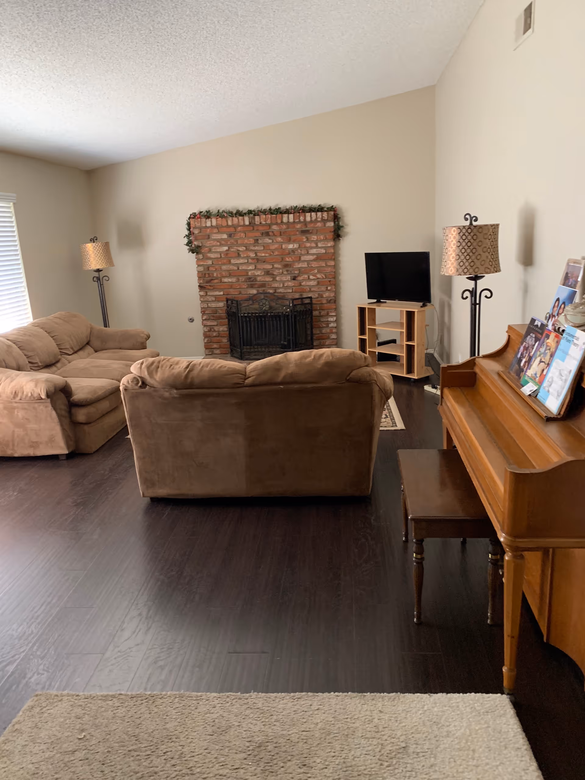 Cozy living room with brown couches, brick fireplace, piano, and TV in a common area.