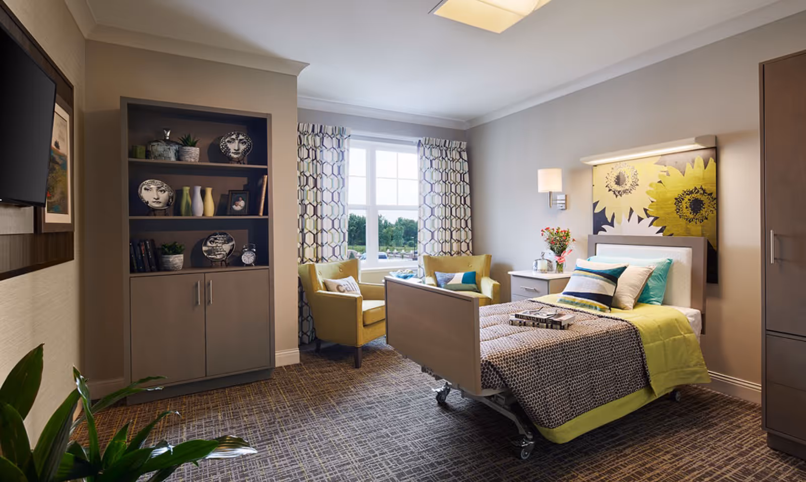 A well-lit bedroom in a senior living facility featuring a single bed with patterned bedding and multiple pillows. Behind the bed is a large artwork of yellow sunflowers. To the right of the bed is a nightstand with a lamp and a vase of flowers. Two yellow armchairs are placed near a window with patterned curtains, offering a view of greenery outside. A built-in shelving unit with decorative plates, vases, books, and plants is on the left wall. A flat-screen TV is mounted on the wall adjacent to the shelving unit. The room has a carpeted floor and neutral-colored walls.