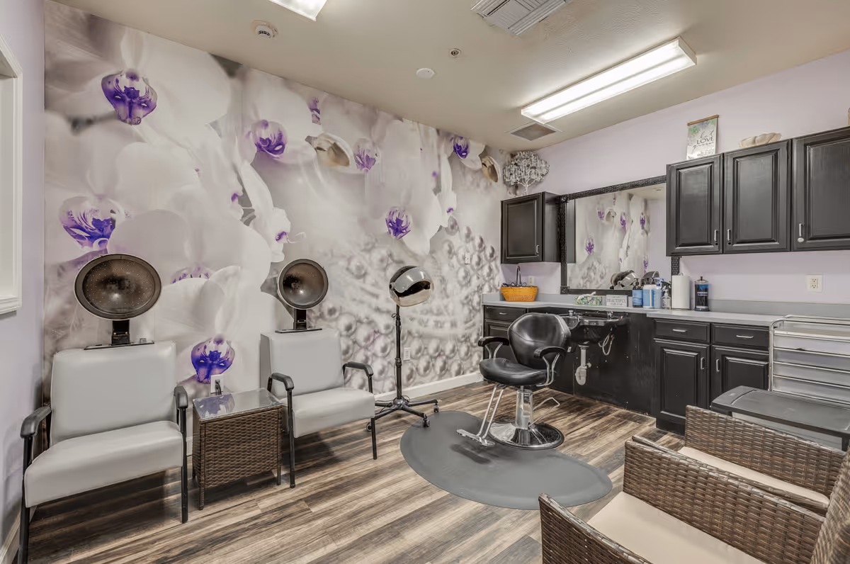 Interior of a salon room with two gray chairs under hair dryers, a black salon chair in front of a large mirror, black cabinets, and a wall decorated with a large floral mural featuring white and purple flowers.