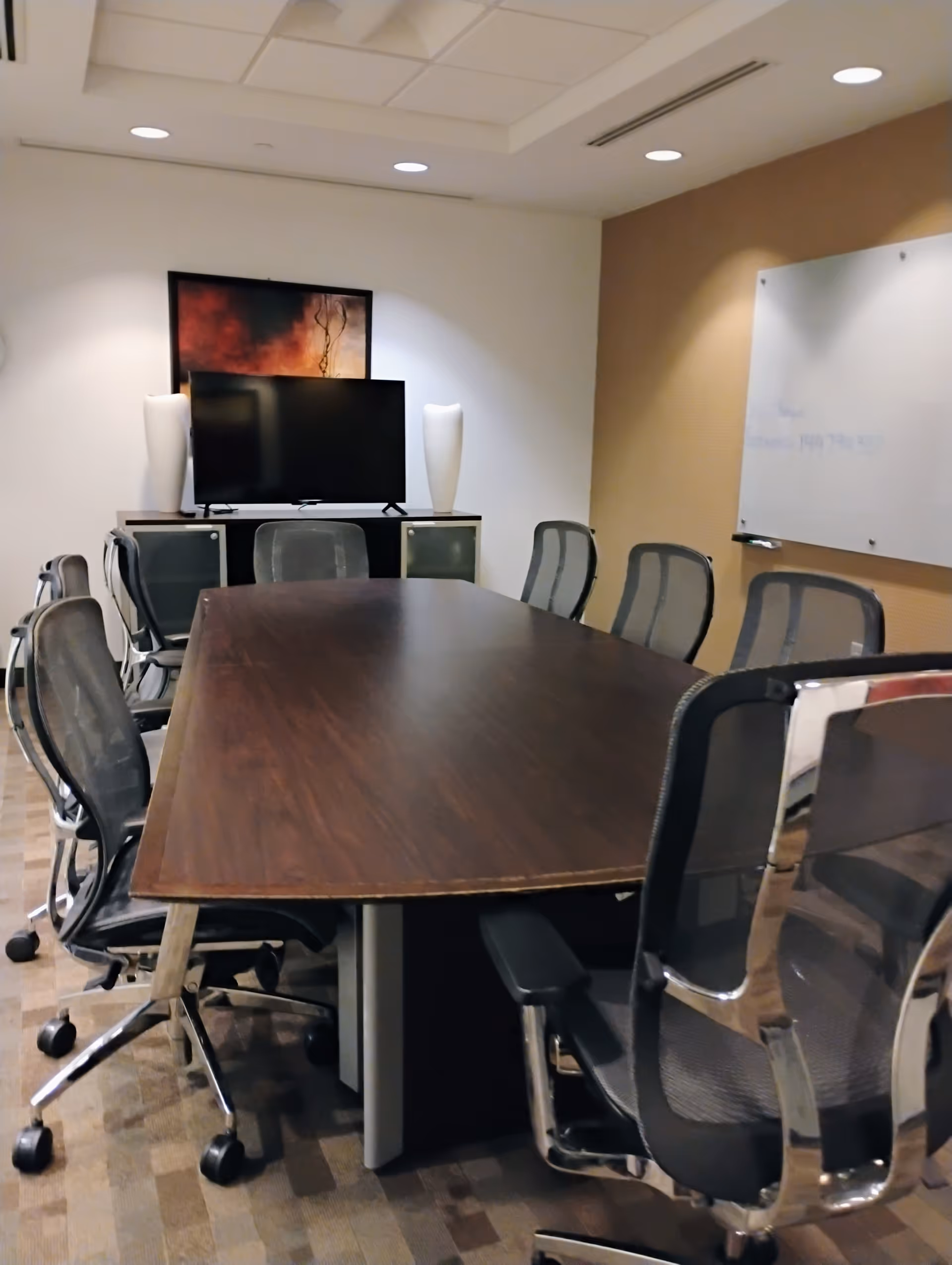 A modern conference room with a long wooden table, office chairs, TV, and whiteboard.