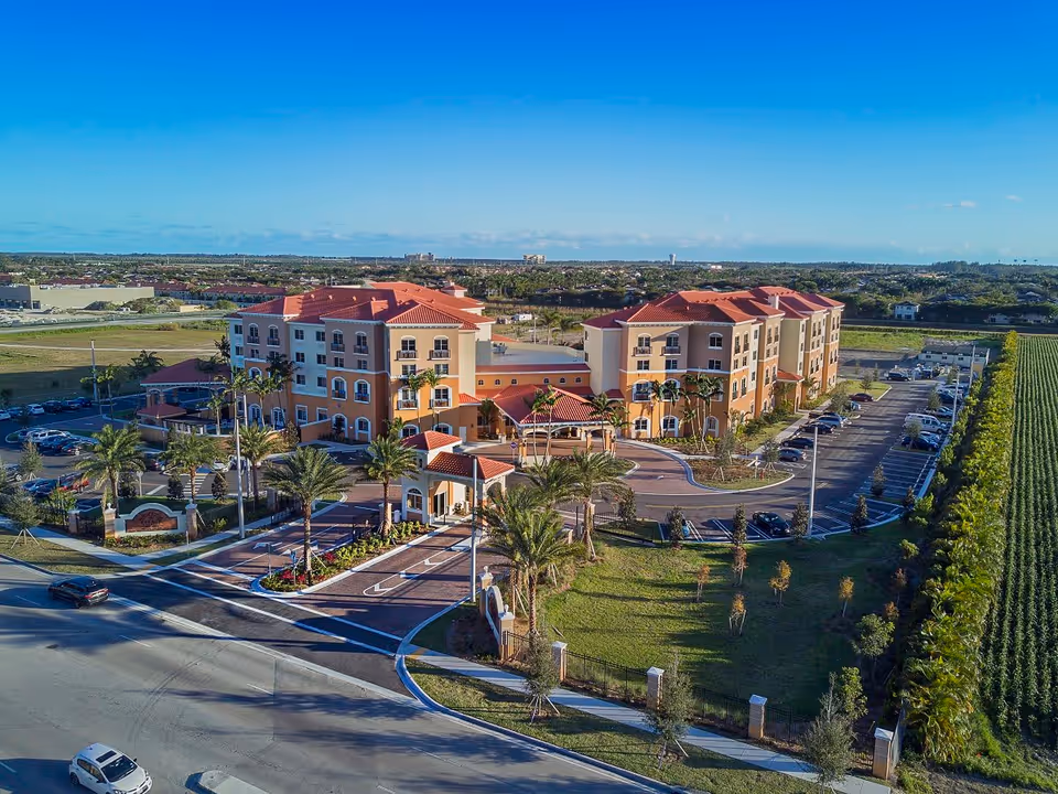 Aerial view of The Palace Gardens assisted living community showcasing the building's facade, entrance, and surrounding landscaping with palm trees.
