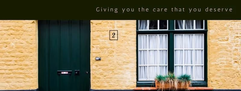 Front of a yellow brick building with a green door, green-framed windows, and potted grasses.