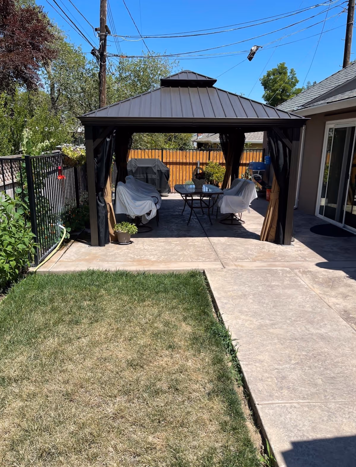 Backyard patio with a metal-roof gazebo over covered chairs and a table, adjacent concrete walkway and small lawn.