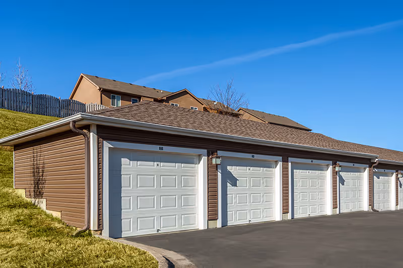 Row of white garage doors in a brown building, with a paved driveway and blue sky.