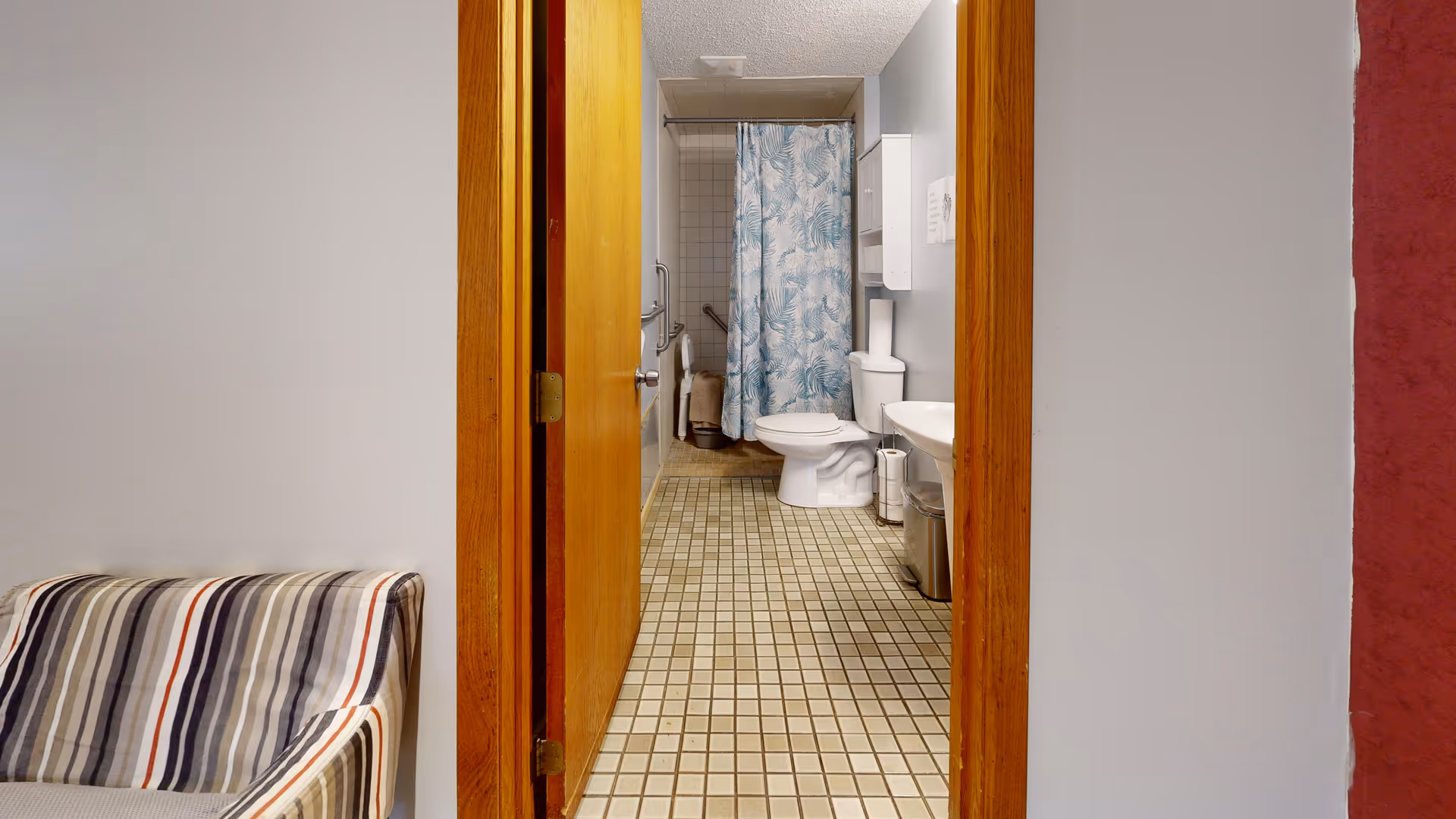Door opens to a bathroom with toilet, sink, and blue patterned curtain; an armchair sits outside.