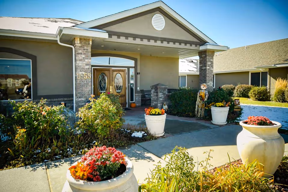 Entrance of a senior living building with double wooden doors, brick columns, potted flowers, and autumn decorations.