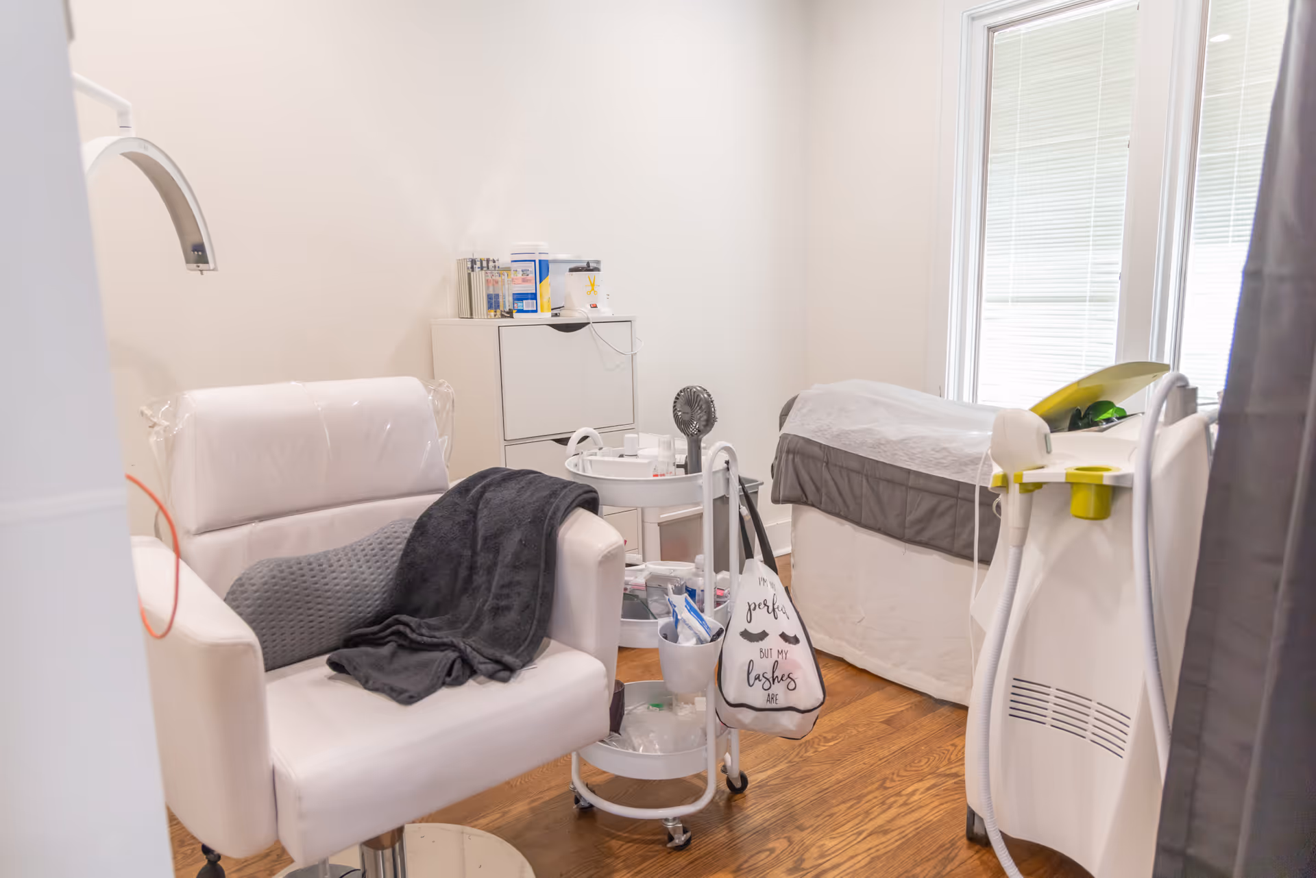 An interior treatment room with a white chair, examination bed, cart, and medical supplies.