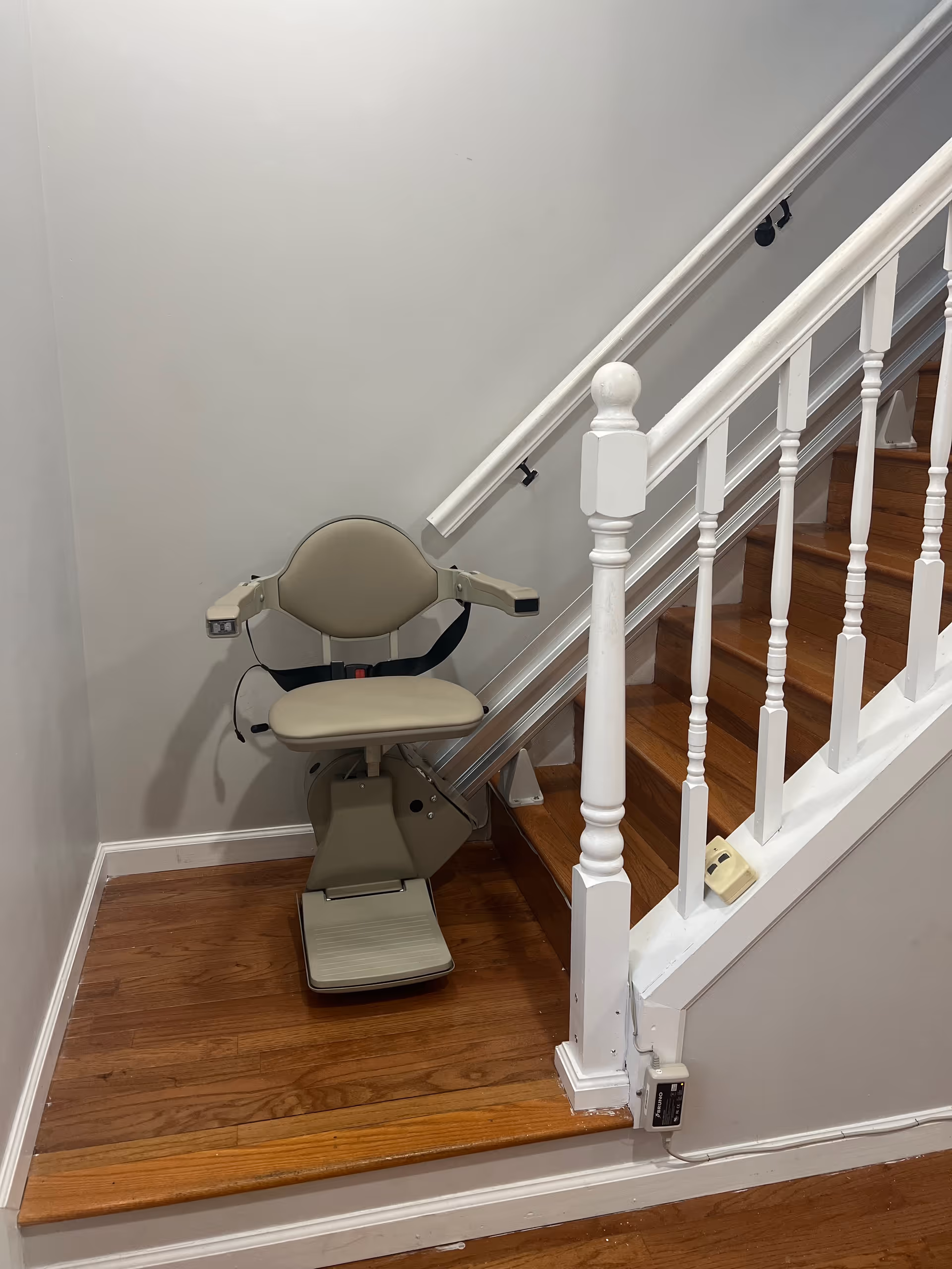 A beige stair lift chair installed beside a wooden staircase in a facility hallway.