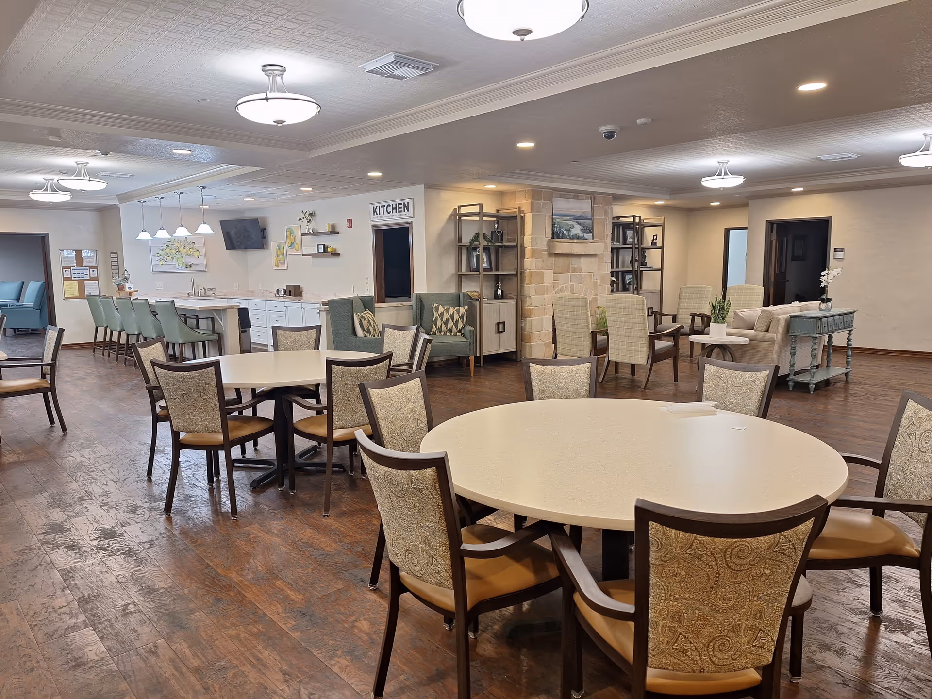 Spacious senior living dining area with round tables, chairs, and a kitchen in the background.