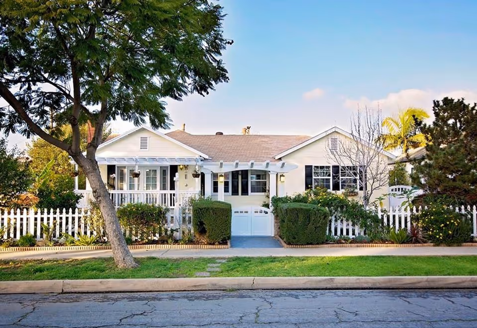 White single-story cottage with a front porch, picket fence, garage door, and landscaped yard under a large tree.