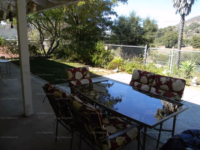 Outdoor patio with glass table and floral cushions overlooking a fenced yard.