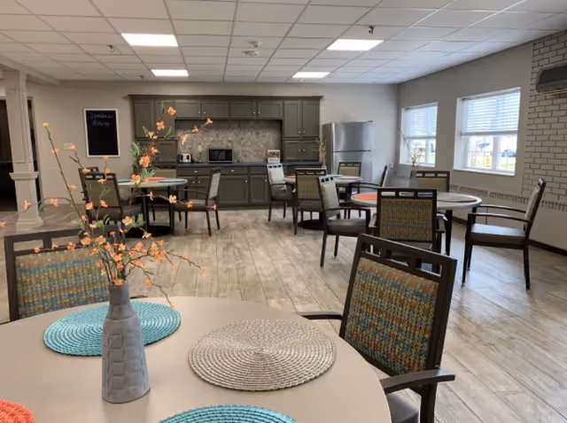 Communal dining area with round tables and chairs, placemats and a vase of flowers, with a kitchenette and refrigerator in the background.