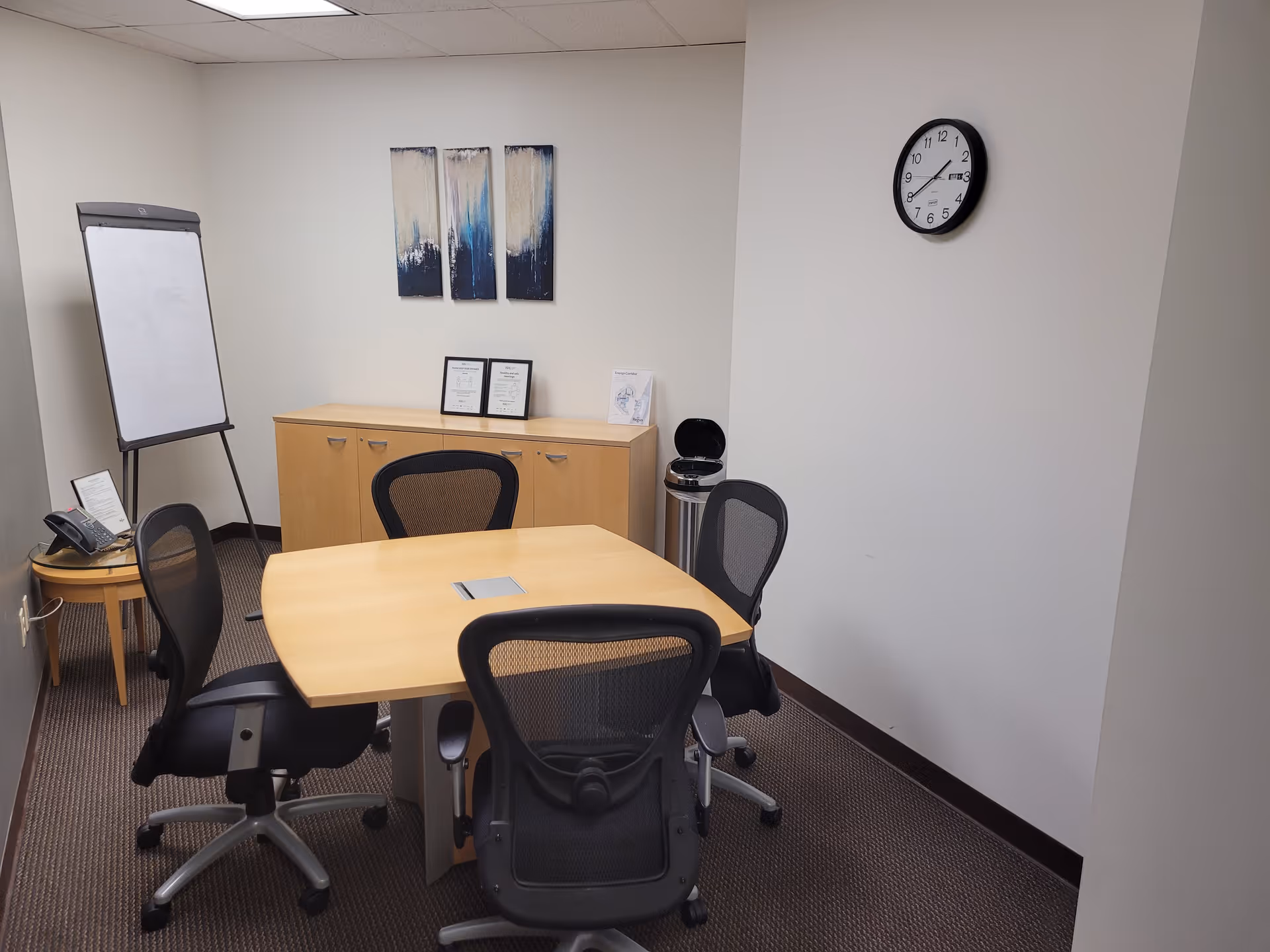 A small conference room with a wooden table, office chairs, a whiteboard, and a wall clock.
