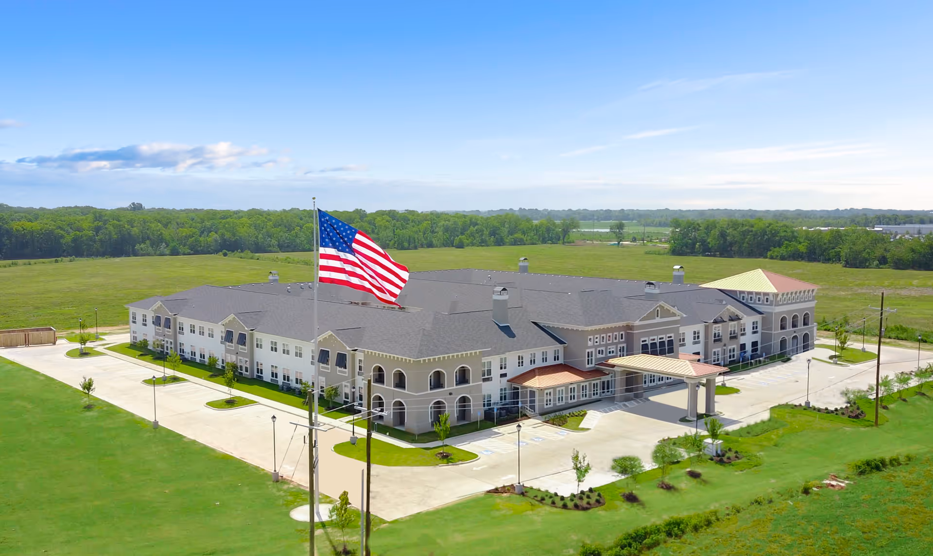 Aerial view of The Blake at Bossier City, a large two-story senior living facility building with a gray roof and beige exterior. The building is surrounded by green lawns and a parking area. An American flag on a tall flagpole is prominently visible in the foreground. The background shows open fields and a partly cloudy sky.