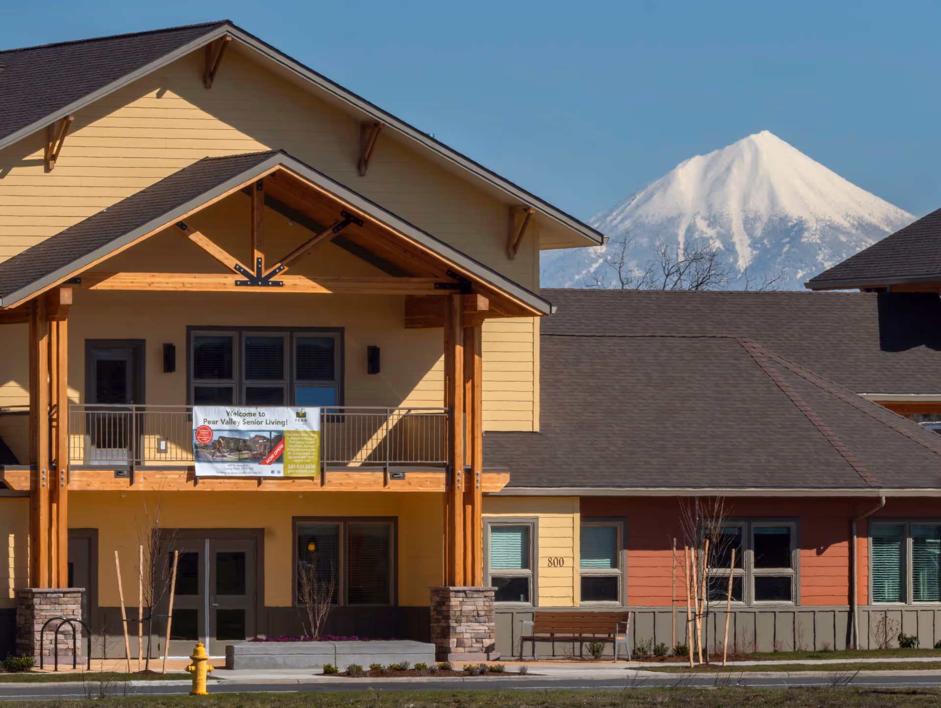 Front exterior of Pear Valley Senior Living building with a balcony banner and a snow-capped mountain in the background.