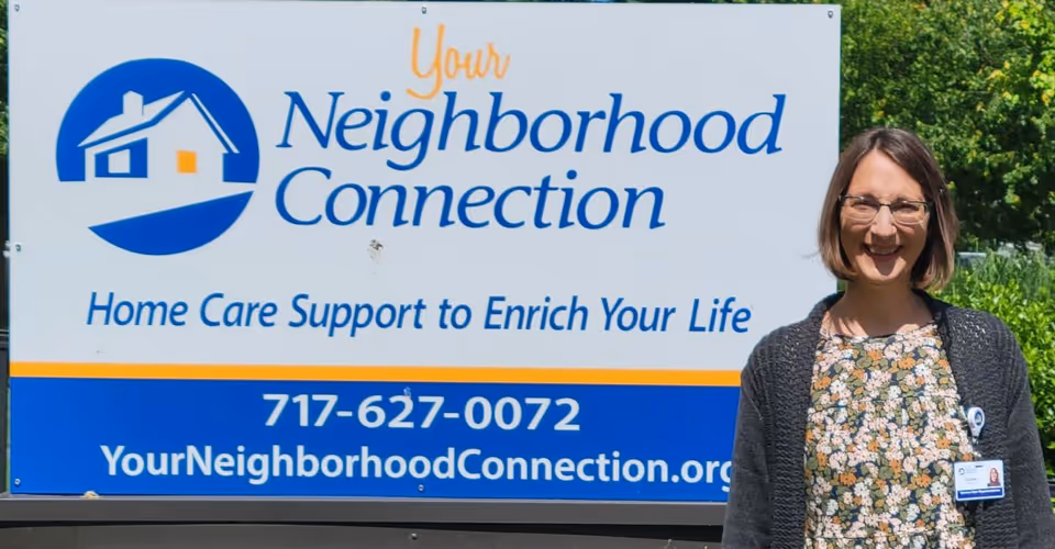 Smiling woman stands beside a large Your Neighborhood Connection sign outdoors.
