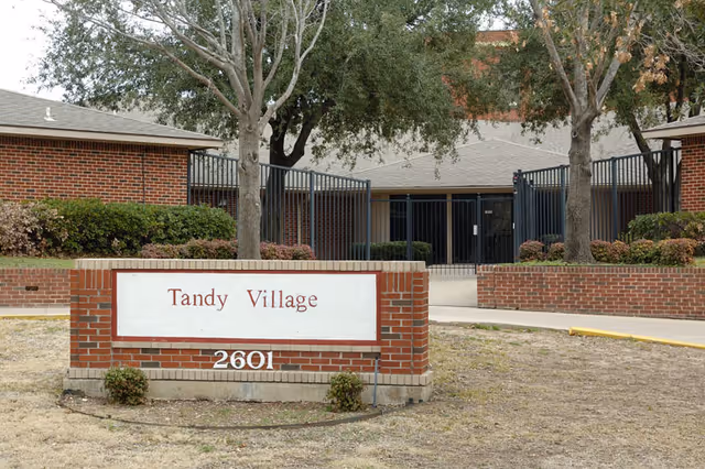 Brick sign reading "Tandy Village" with address "2601" on a lawn in front of a brick building entrance framed by trees.