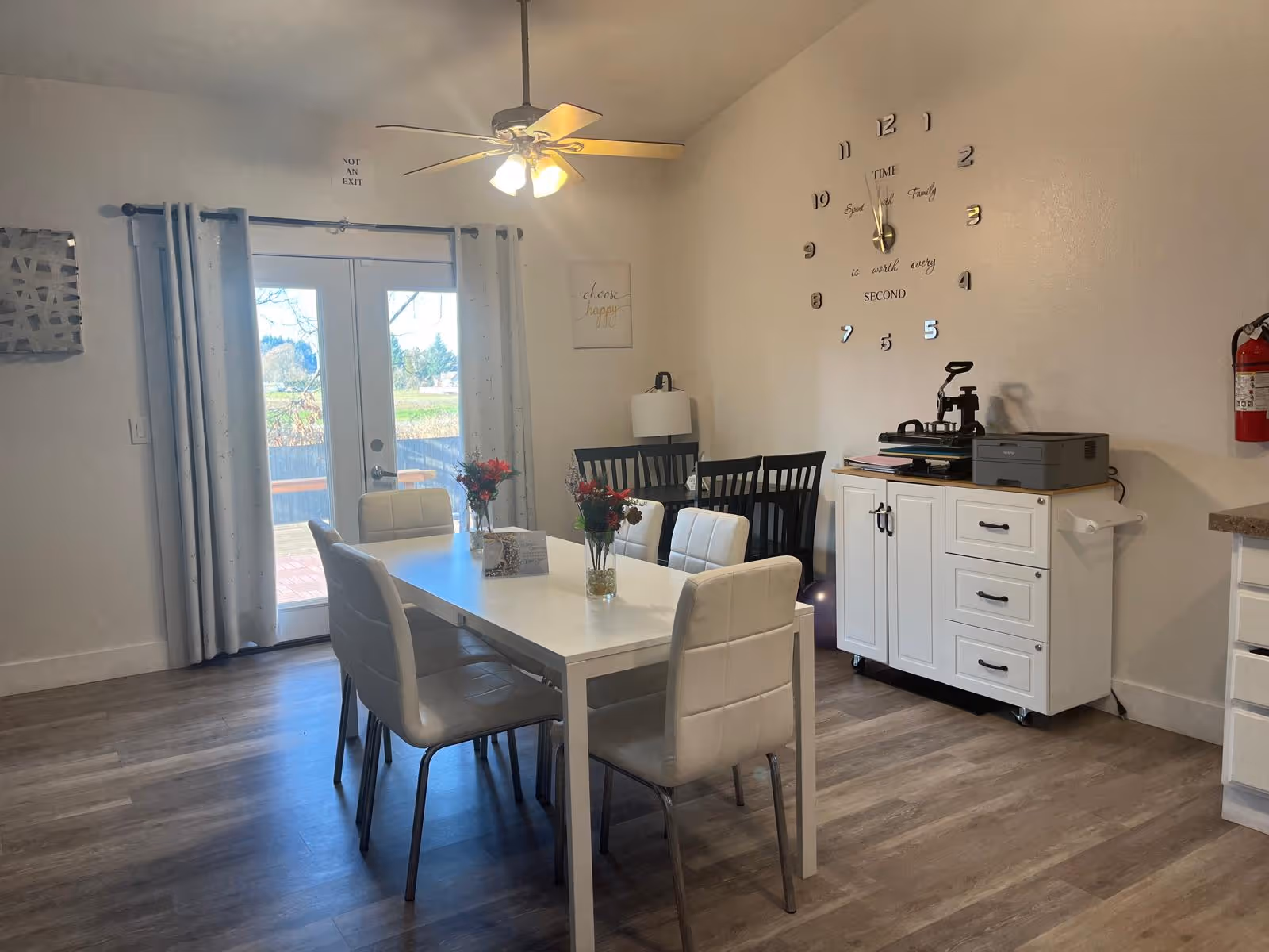 Bright dining room with white table and chairs, wall clock, and printer cabinet.