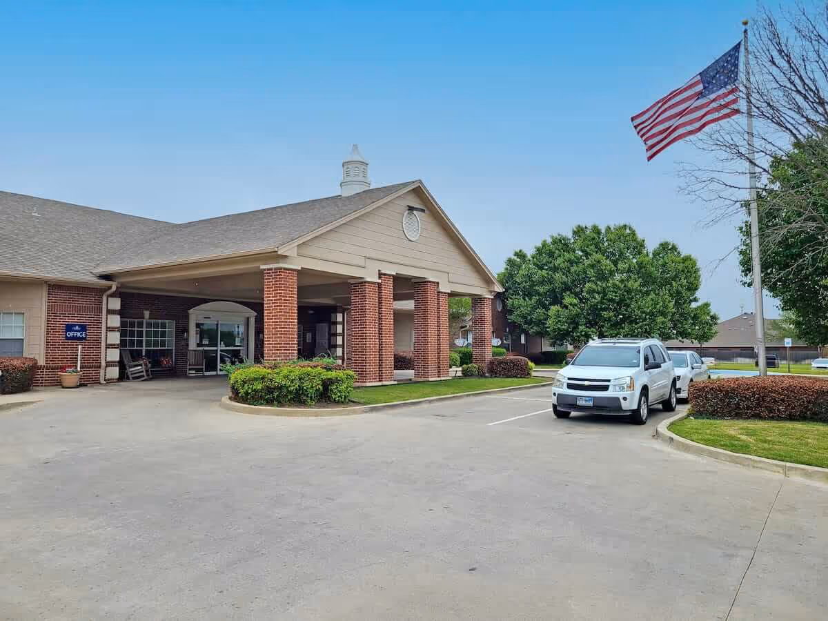 Entrance of a brick senior living building with a covered porte-cochère, parked cars, landscaping, and an American flag.
