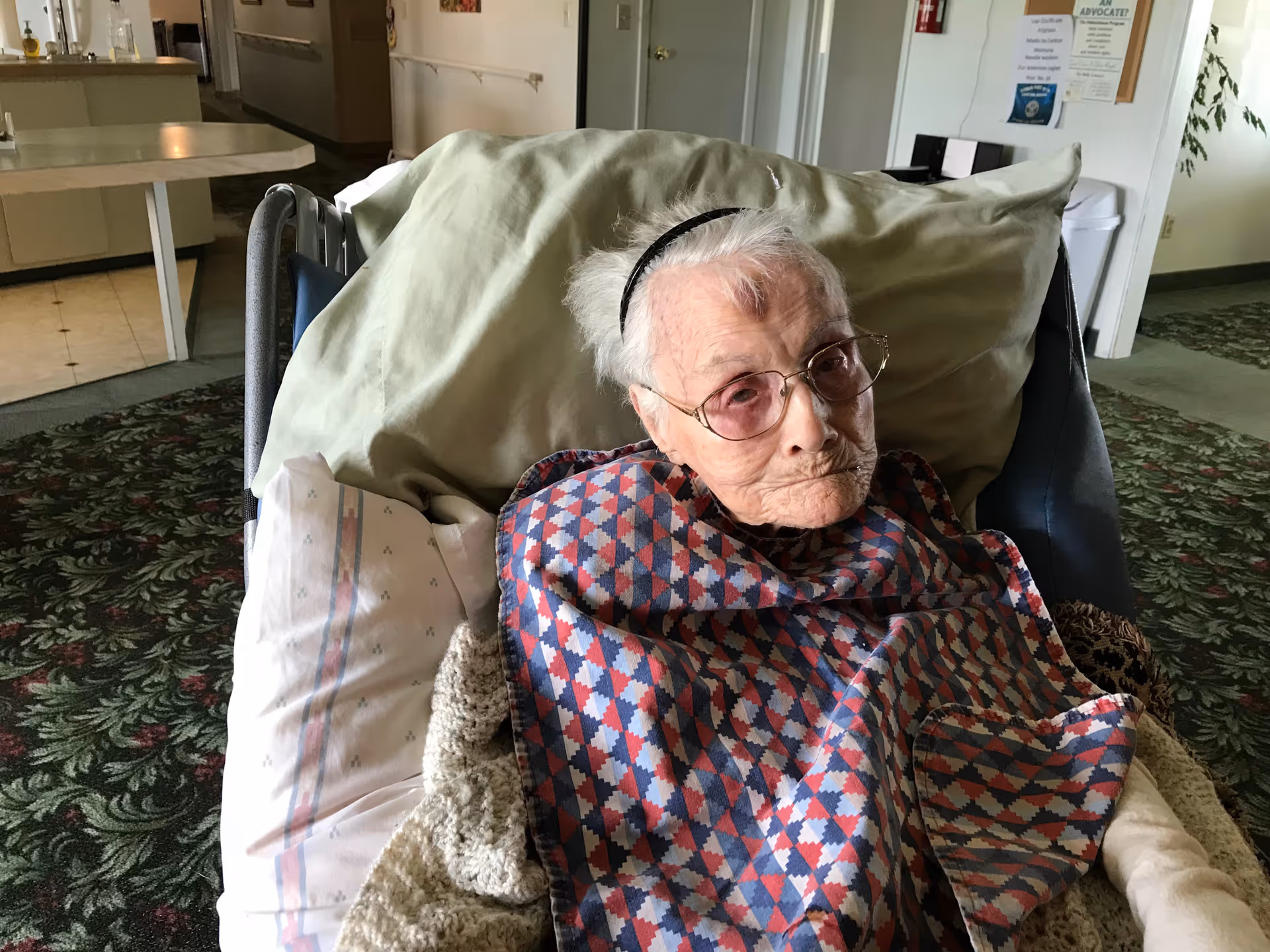 An elderly woman with glasses and a headband is sitting in a wheelchair covered with a patterned blanket. She is indoors in a room with floral carpet and light-colored walls, with a kitchen area visible in the background.