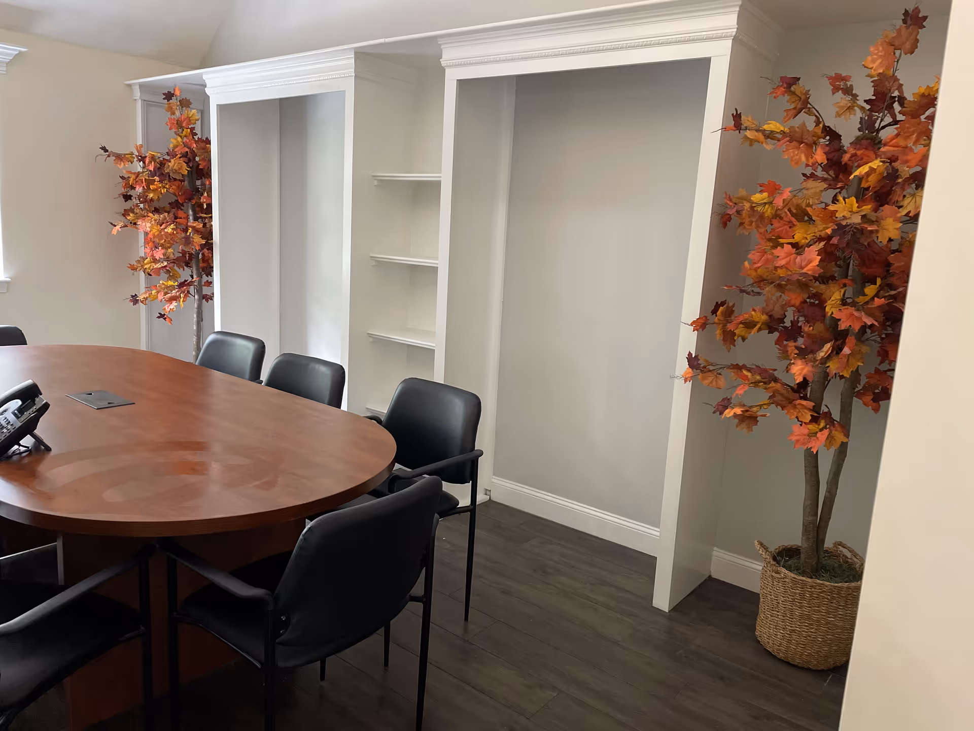 An oval conference table with black chairs, white shelves, and autumn trees in pots.