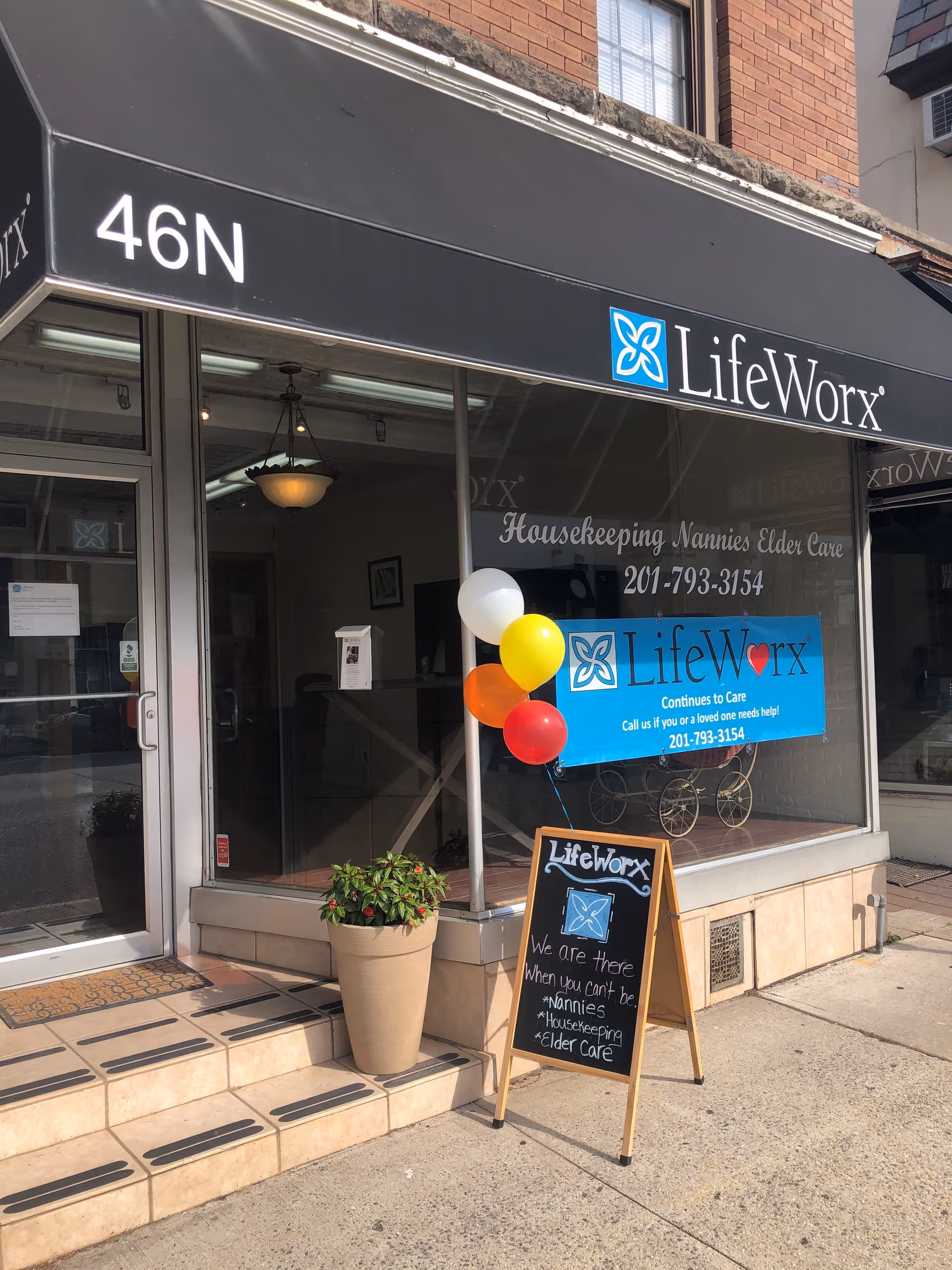 Storefront with LifeWorx signage, glass doors, colorful balloons, a chalkboard sign, and a potted plant outside.