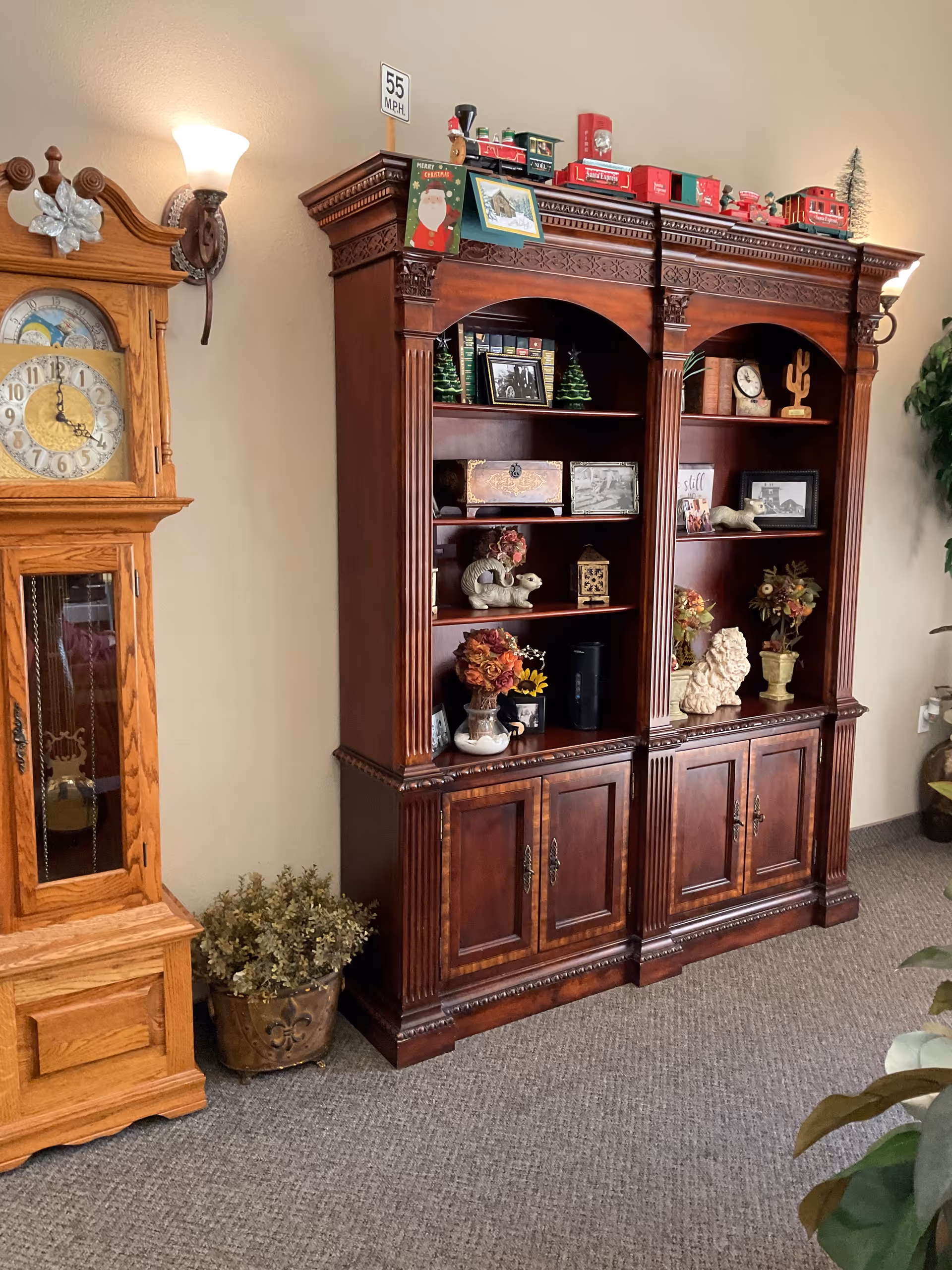 A wooden grandfather clock and a large wooden bookshelf with decorative carvings. The bookshelf is decorated with various items including small Christmas trees, framed photos, a toy train, and floral arrangements. The room has beige walls, carpeted floor, and wall-mounted light fixtures.