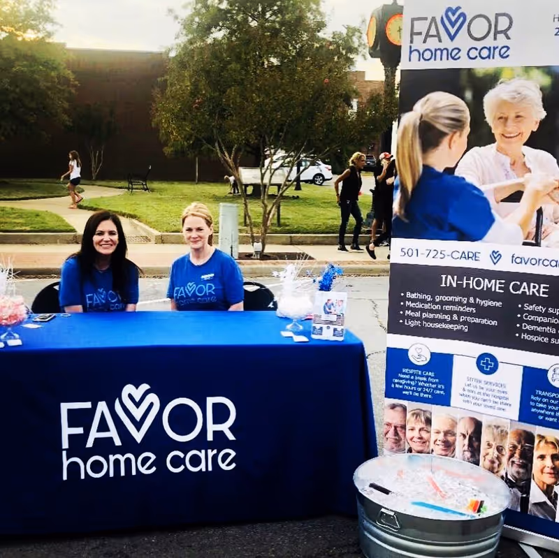 Two staff members sit at a blue table with a Favor Home Care banner outdoors.
