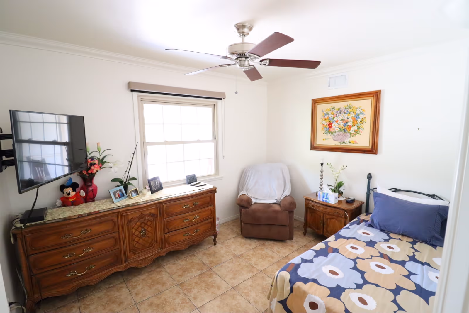 Cozy bedroom with a floral bedspread, dresser, armchair, and TV by a window.