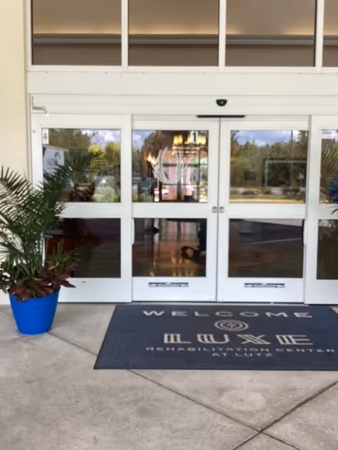 Entrance to Lutz Healthcare & Rehabilitation with glass doors, welcome mat, and potted plants.