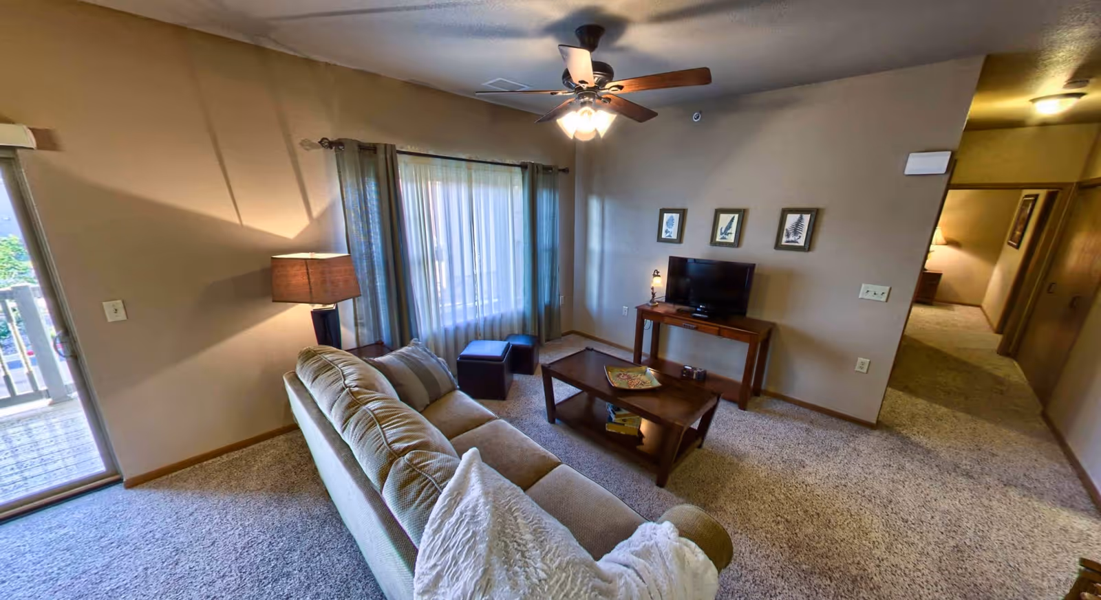 A cozy living room with a beige sofa, a wooden coffee table with books and a decorative plate, a TV on a wooden stand, three framed pictures on the wall, a floor lamp, and a ceiling fan with lights. There is a window with sheer curtains and a sliding glass door leading to a balcony.