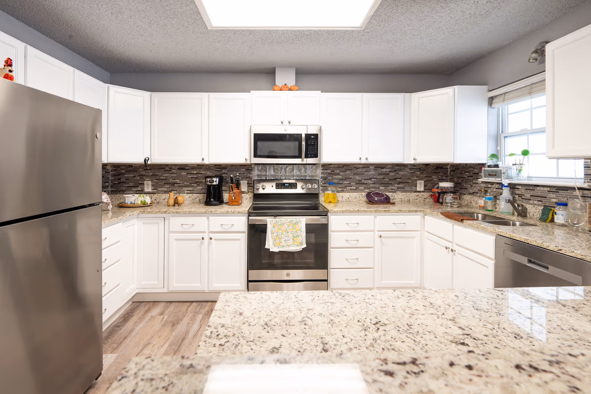 A modern kitchen with white cabinets, granite countertops, and stainless steel appliances including a refrigerator, stove, microwave, and dishwasher. There is a window above the sink letting in natural light, and various kitchen items such as a coffee maker, knife block, and dish soap are visible on the counters.