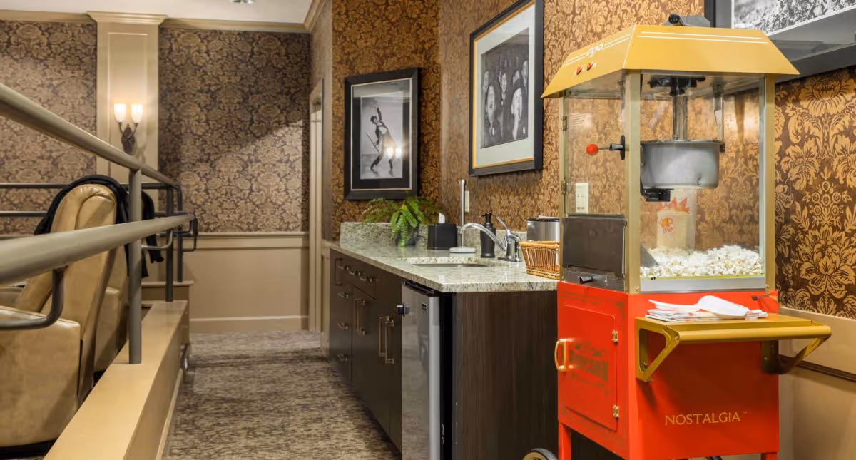 Small interior lounge area with a popcorn machine next to a granite-topped kitchenette and theater-style seating against patterned wallpaper.