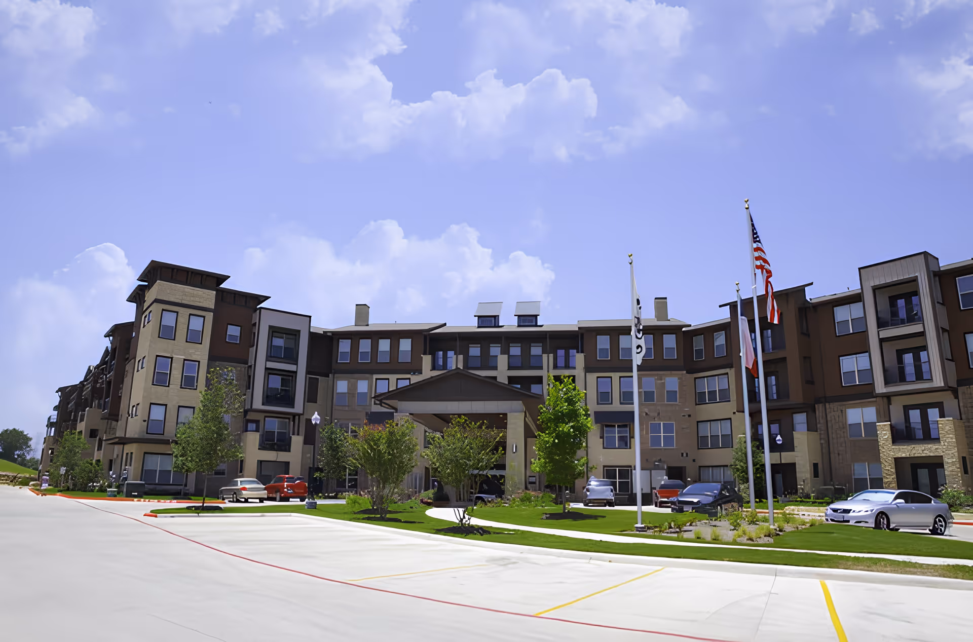 Exterior view of a multi-story senior living facility building with a covered entrance, several parked cars, green landscaped areas with trees and shrubs, and three flagpoles displaying flags under a partly cloudy sky.
