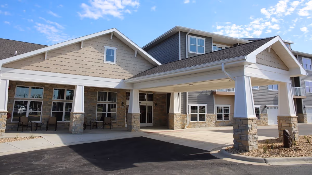 Exterior view of a senior living facility building with a covered entrance supported by stone and white pillars. The building has multiple windows, beige and gray siding, and a paved driveway. There are chairs and small tables arranged outside near the windows.
