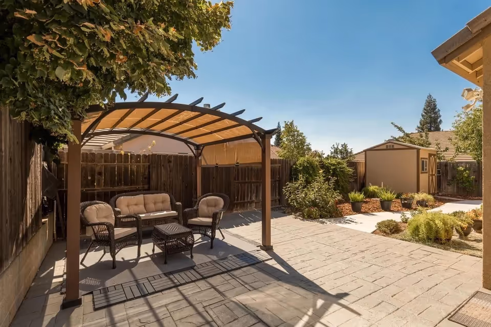 Outdoor patio with wicker seating under a curved canopy, fenced yard, and garden planters.