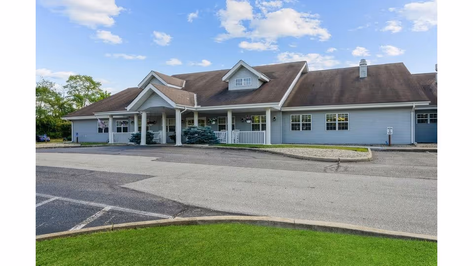 Exterior view of a single-story senior living facility building with a covered entrance, light blue siding, multiple windows, and a sloped roof under a partly cloudy sky. There is a paved driveway and parking area in front, with some greenery and trees in the background.