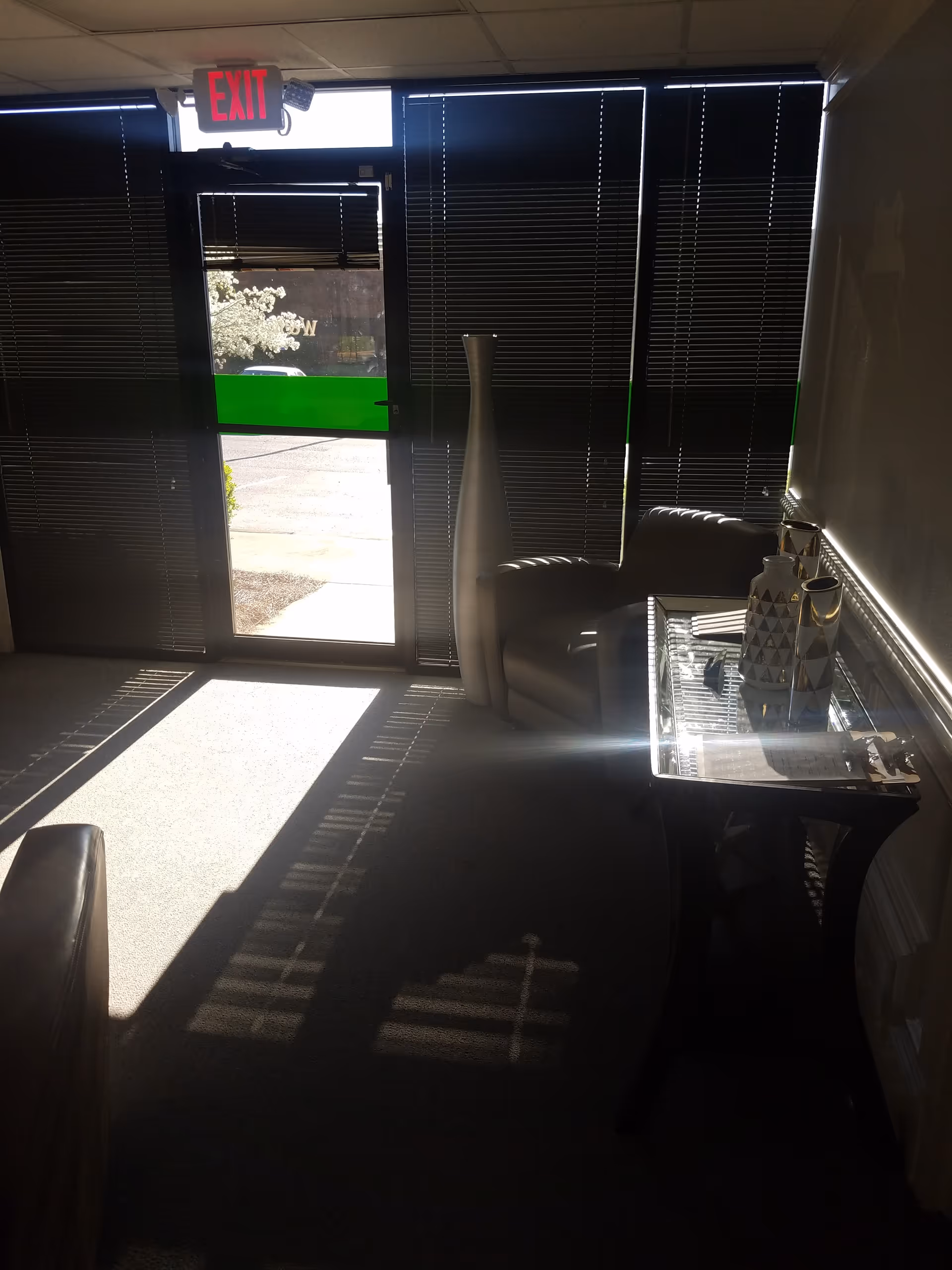 Sunlight streams through blinds into a small interior lobby with couch, glass table, and exit sign.